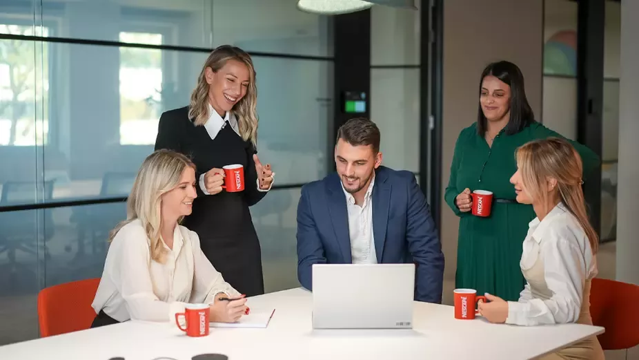 Zaposleni okupljeni oko laptopa tokom sastanka u modernoj kancelariji, držeći NESCAFÉ šolje.